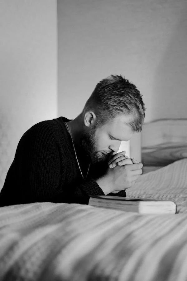 A man prays to God while kneeling in bed, focusing closely. 